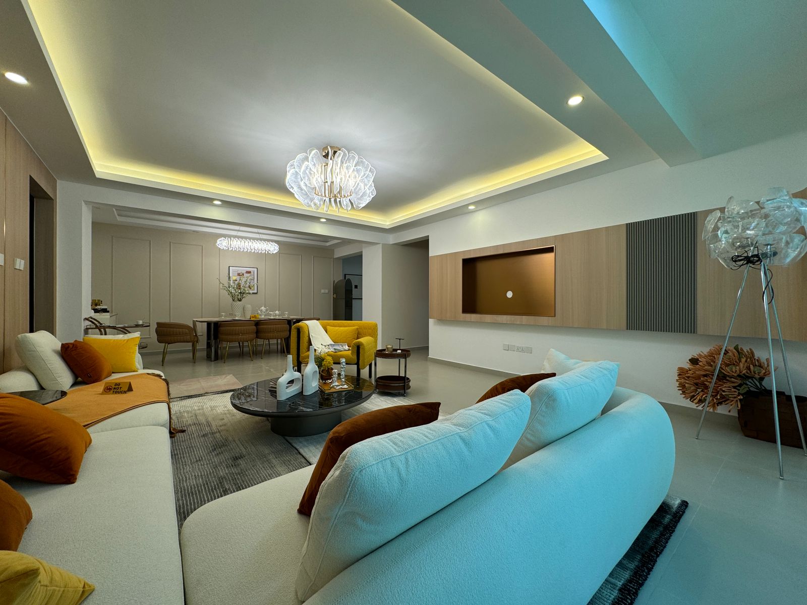 Royal Legend Residence spacious living room featuring cream sofas, yellow accent chair, black coffee table, decorative chandelier, and modern TV wall.