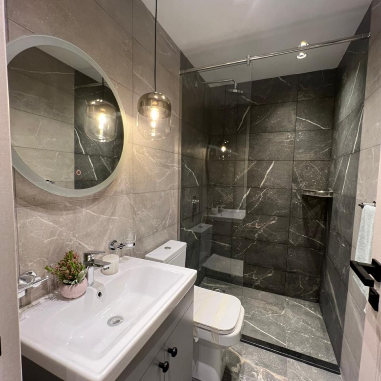 Symphony Residence modern bathroom featuring a glass-enclosed shower, marble-effect wall tiles, vanity sink, round mirror and toilet.
