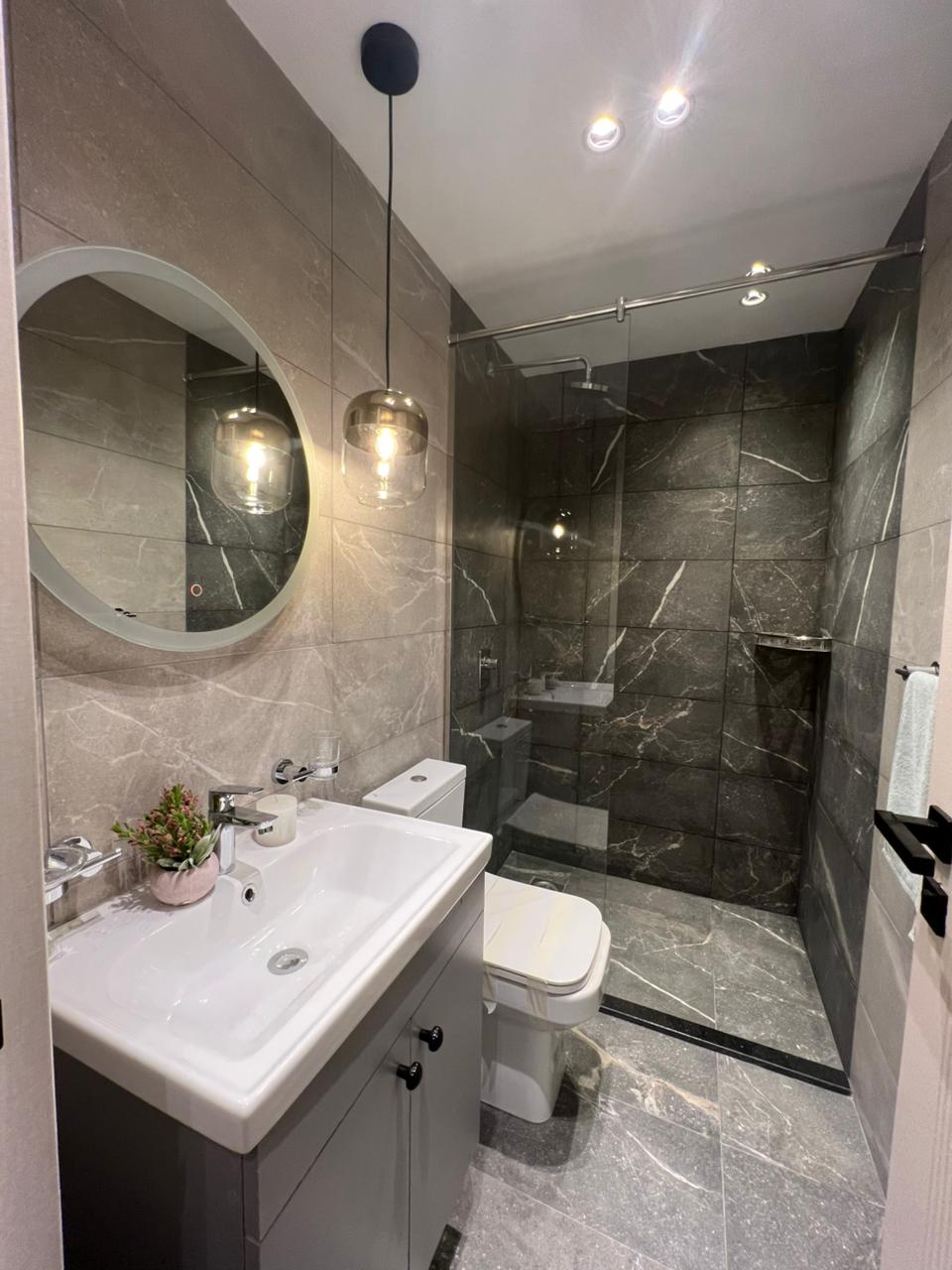 Symphony Residence modern bathroom featuring a glass-enclosed shower, marble-effect wall tiles, vanity sink, round mirror and toilet.