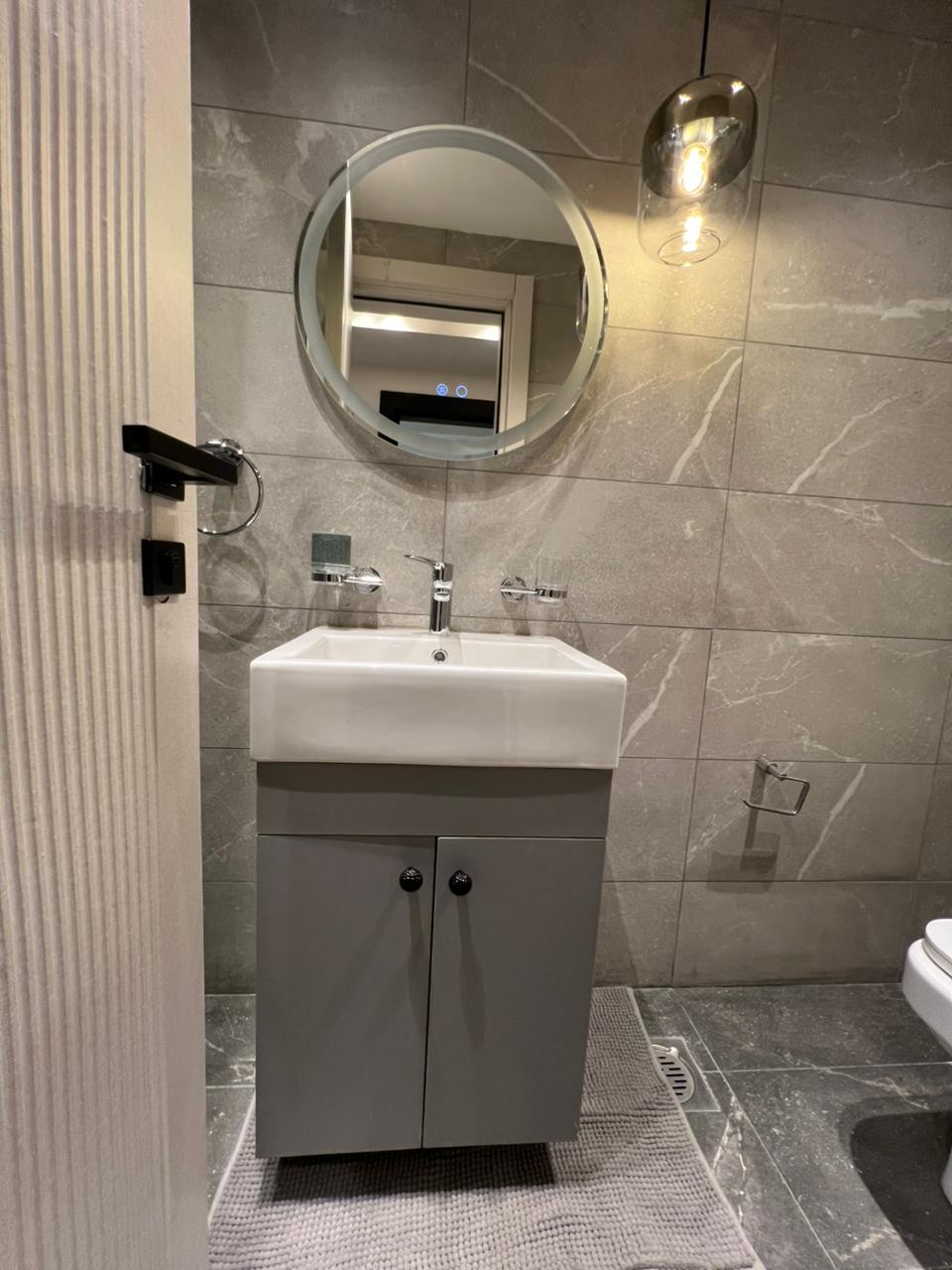 Symphony Residence bathroom vanity featuring a white ceramic washbasin, grey storage cabinet, round mirror and decorative pendant light.