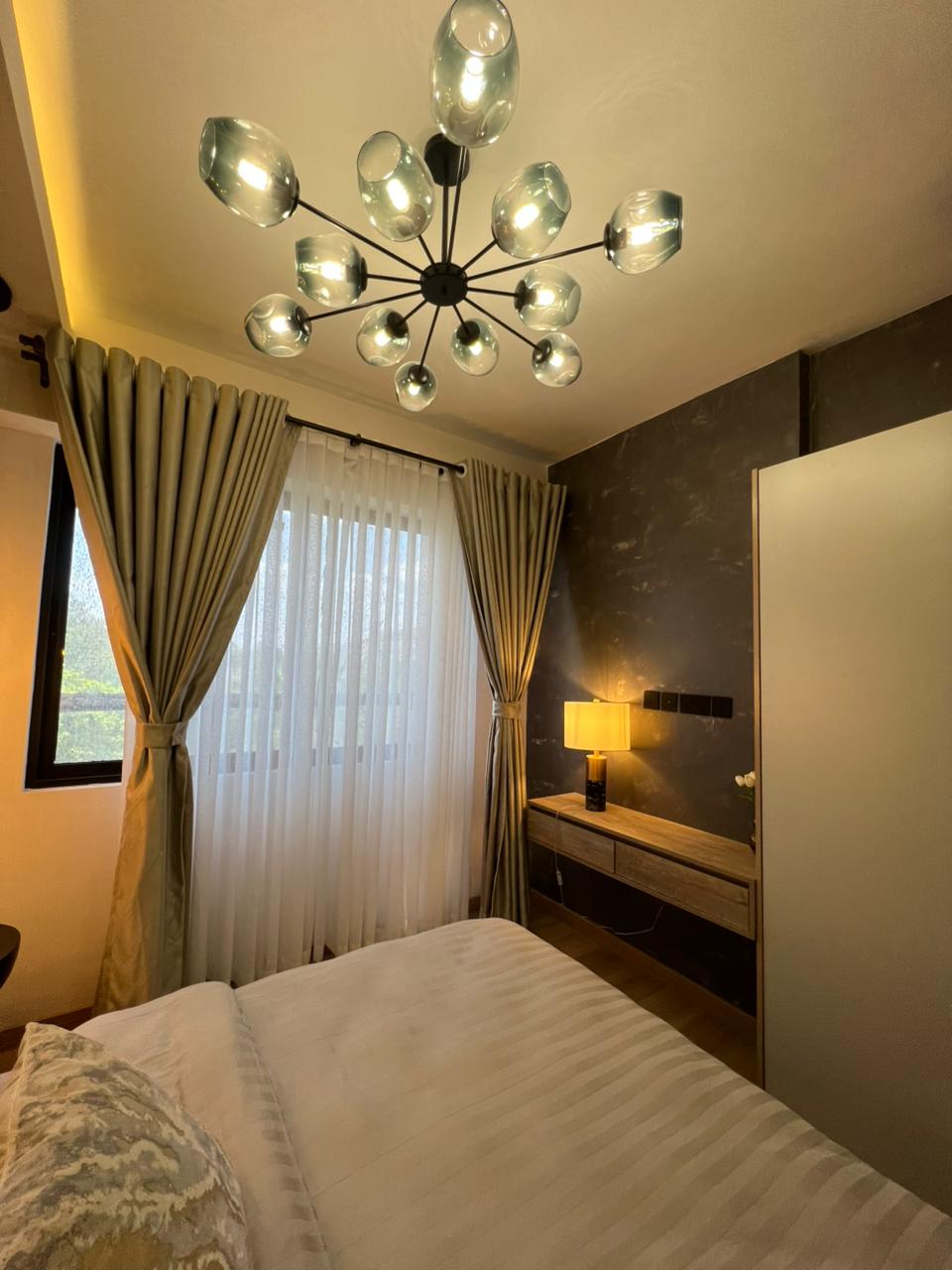 Symphony Residence bedroom interior featuring a modern glass chandelier, floor-length curtains, wooden dressing table, wardrobe and textured accent wall.
