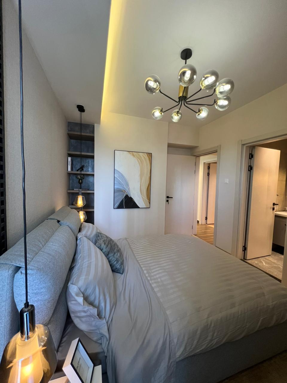 Symphony Residence bedroom featuring a large upholstered bed, pendant lighting, decorative ceiling chandelier, modern wall art and ensuite bathroom access.