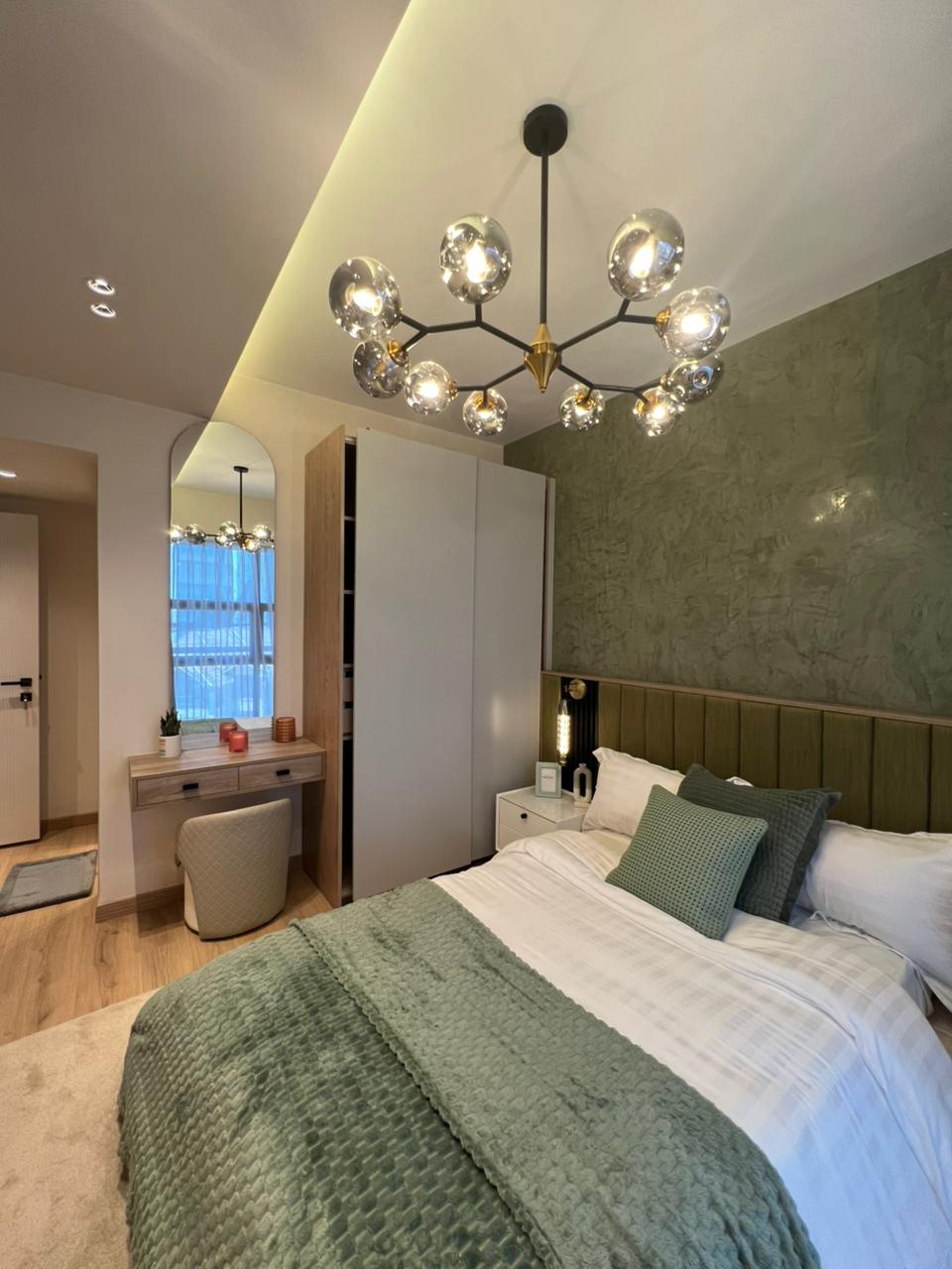 Symphony Residence bedroom featuring a green textured accent wall, upholstered bed, modern wardrobe, dressing table and decorative glass chandelier.
