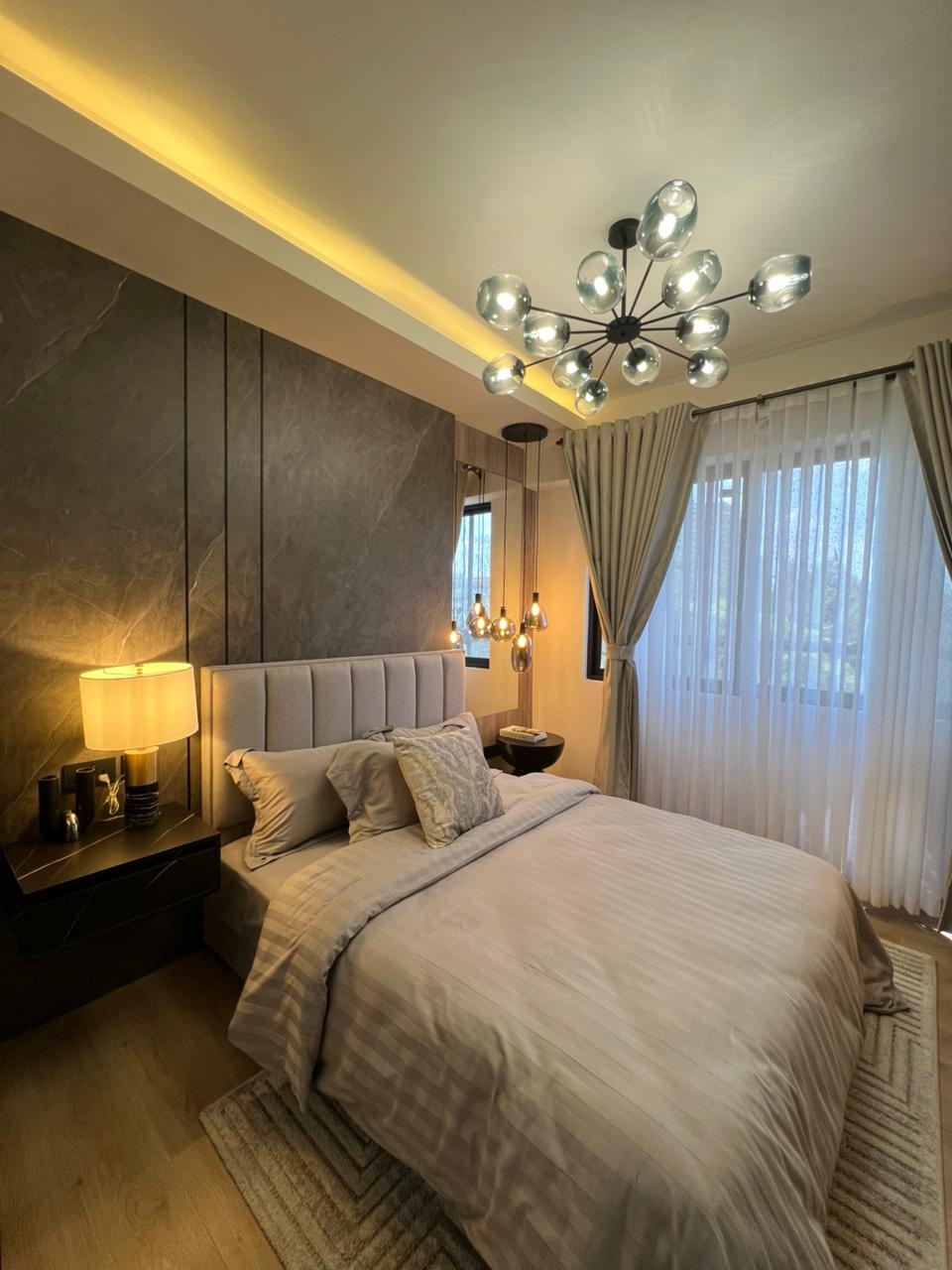 Symphony Residence modern bedroom with a grey upholstered headboard, marble-effect feature wall, glass chandelier, pendant lights and large window.