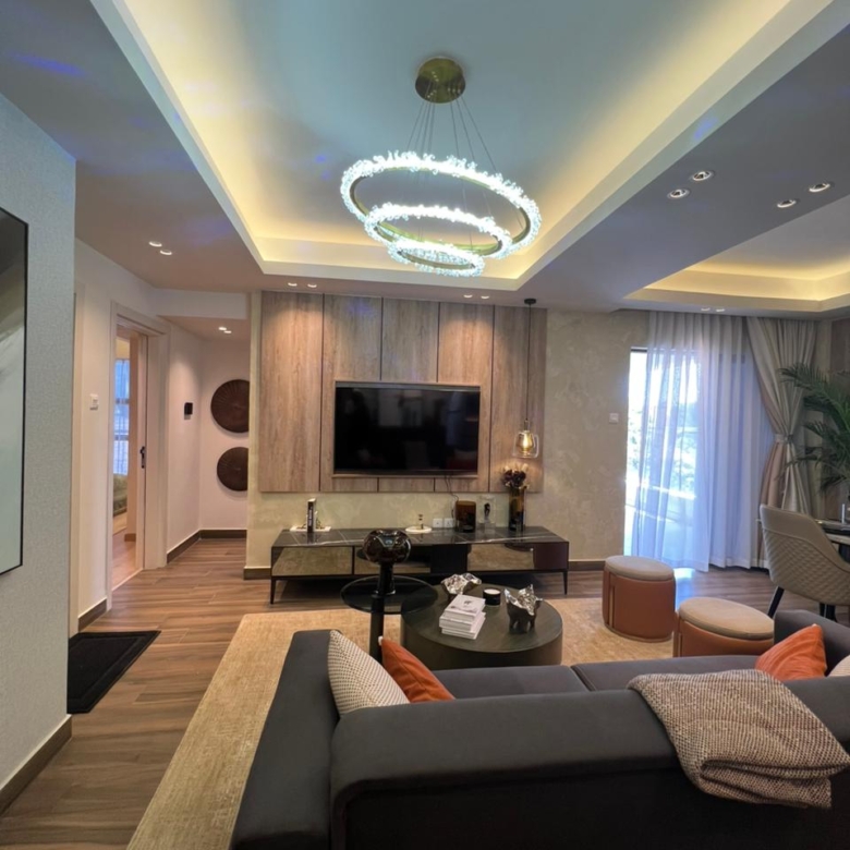 Symphony Residence spacious living room featuring a modern chandelier, wall-mounted TV, grey sectional sofa, round coffee table and large windows.