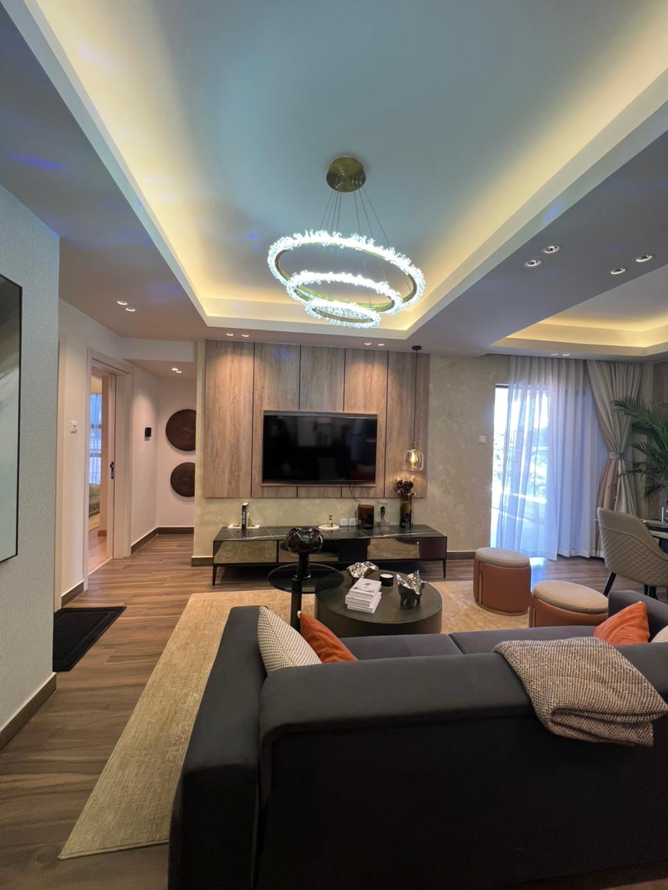 Symphony Residence spacious living room featuring a modern chandelier, wall-mounted TV, grey sectional sofa, round coffee table and large windows.