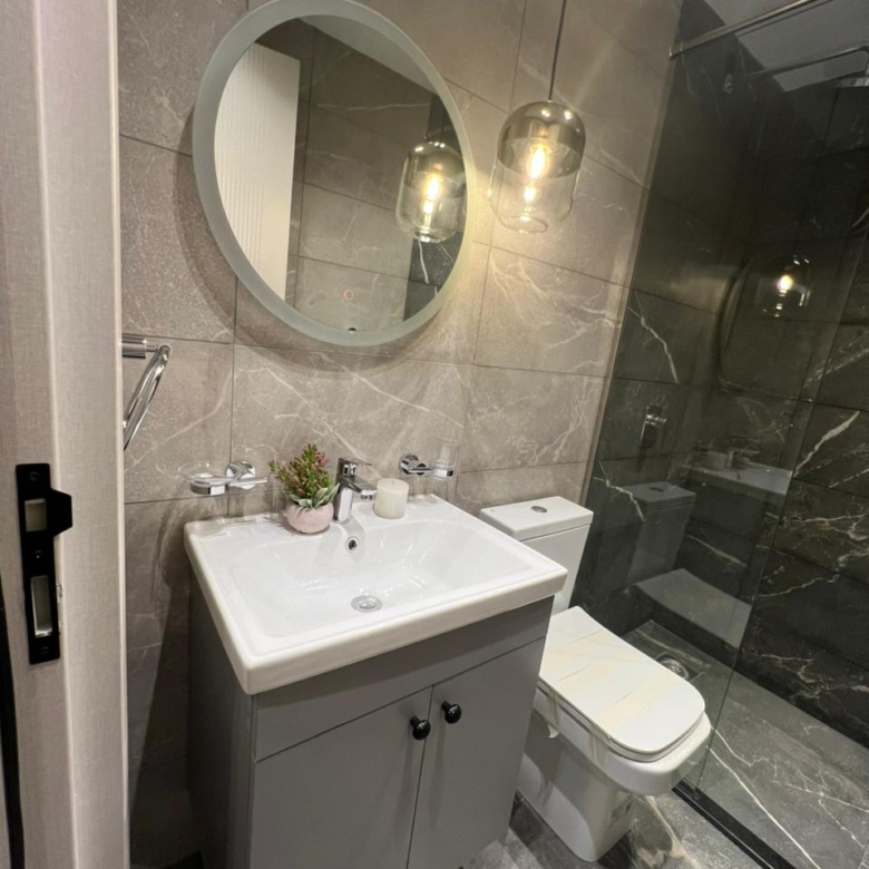 Symphony Residence bathroom sink area featuring a white ceramic washbasin, grey vanity cabinet, round wall mirror and marble-effect tiles.
