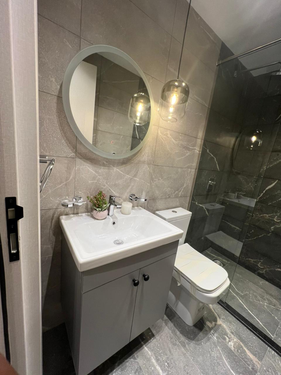 Symphony Residence bathroom sink area featuring a white ceramic washbasin, grey vanity cabinet, round wall mirror and marble-effect tiles.