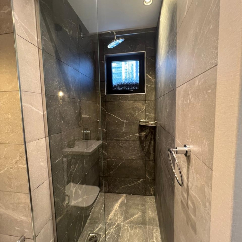 Symphony Residence modern shower enclosure featuring a glass partition, rainfall showerhead, dark marble-effect tiles and bathroom window.