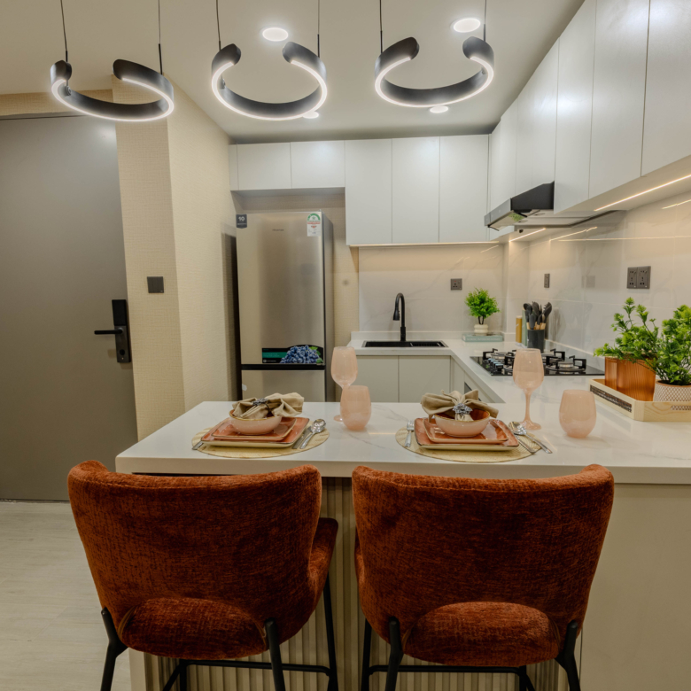 Montbleu apartments 1-bedroom open-plan dining area featuring a glass dining table, upholstered chairs, circular pendant lighting, blue sofa, and modern entertainment wall.