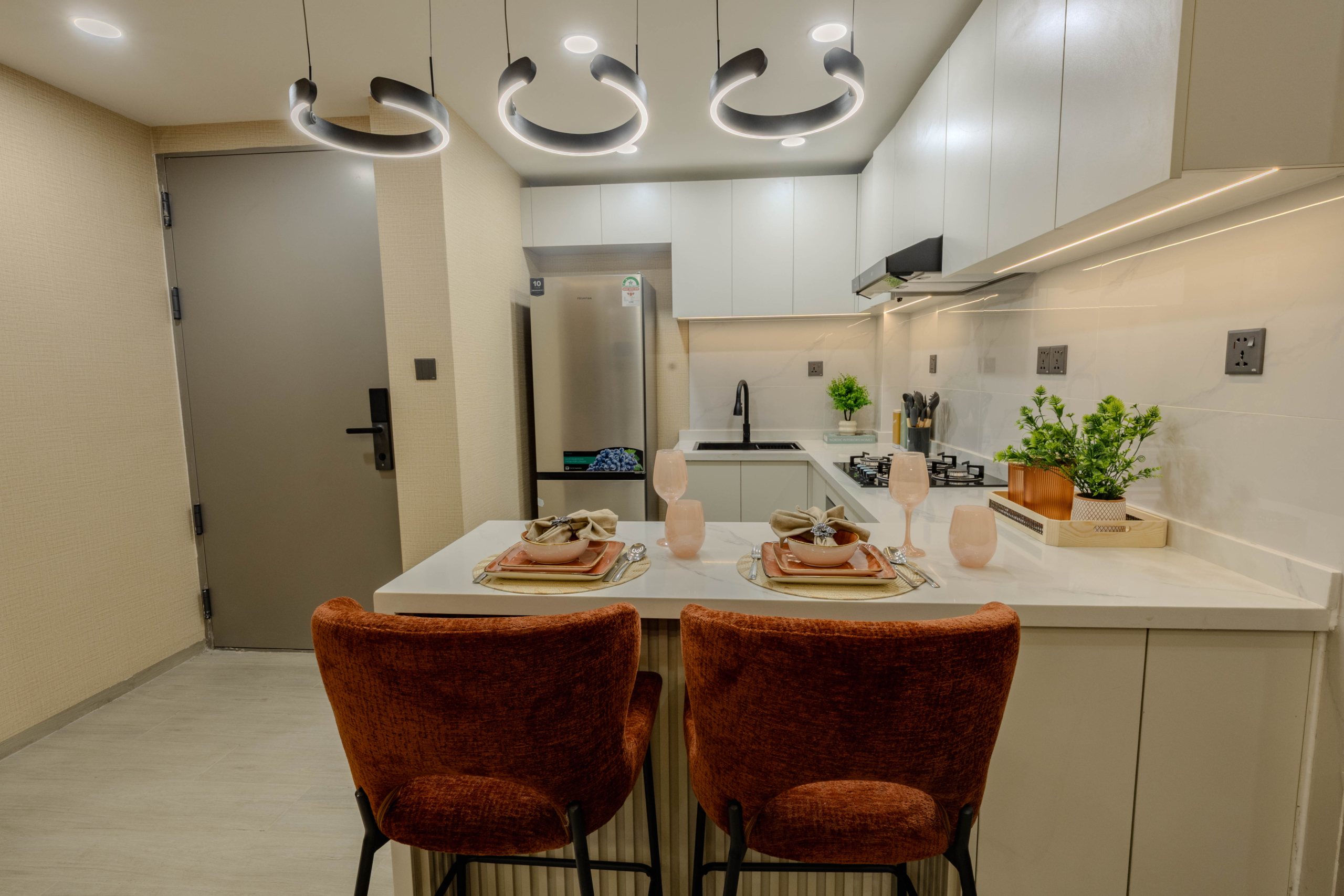 Montbleu apartments 1-bedroom open-plan dining area featuring a glass dining table, upholstered chairs, circular pendant lighting, blue sofa, and modern entertainment wall.