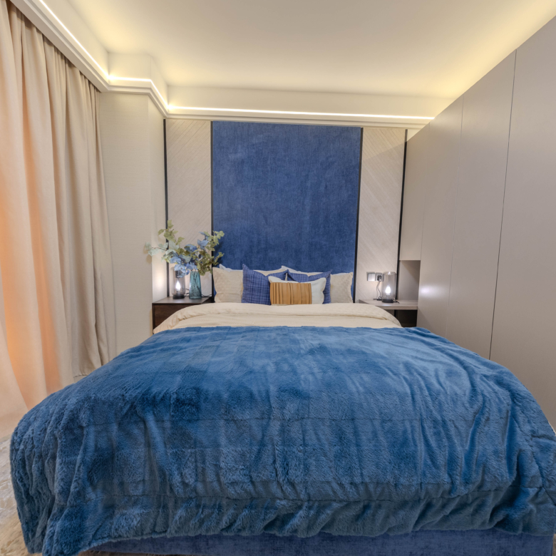 Montbleu apartments 2-bedroom interior showing a stylish bedroom with a blue upholstered headboard wall, comfortable bed, blue bedding, fitted wardrobes, and warm LED ceiling lighting.