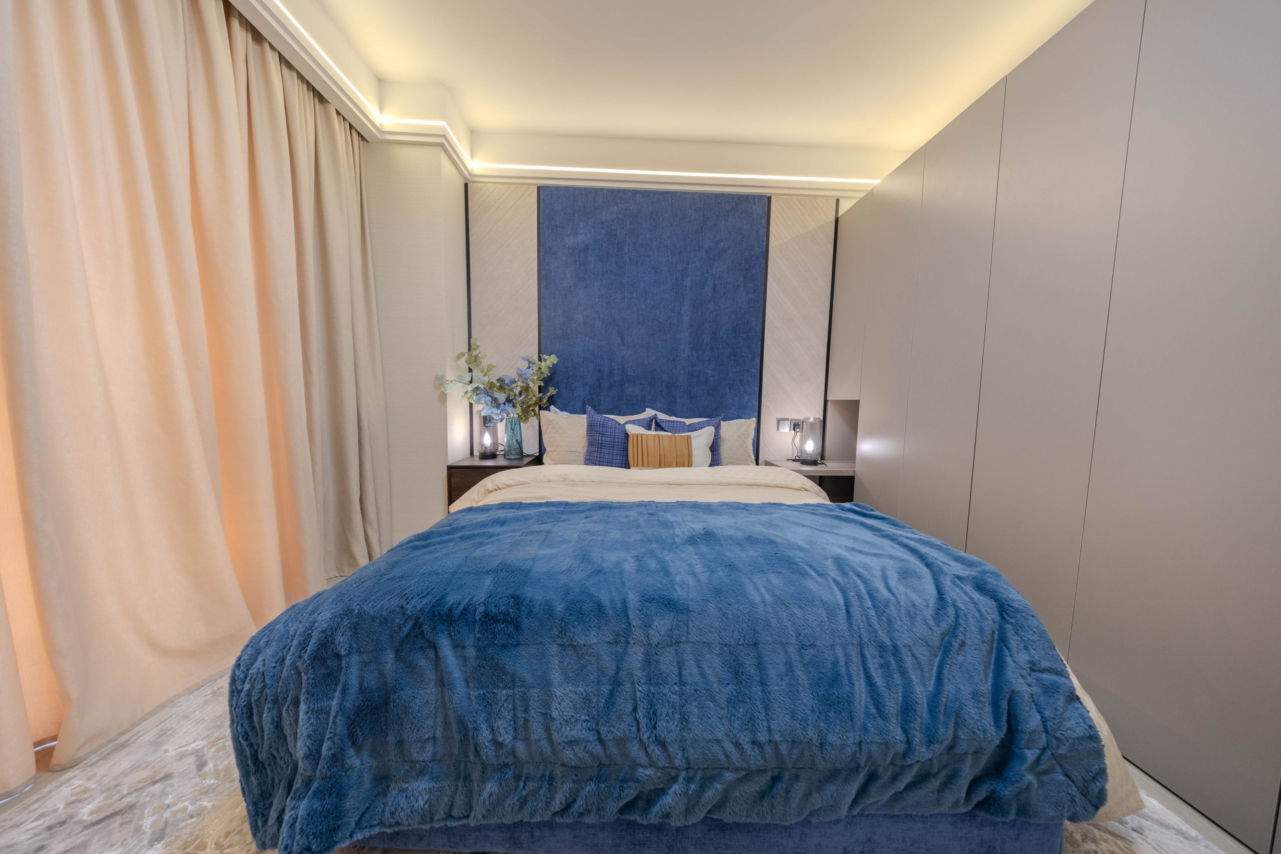 Montbleu apartments 2-bedroom interior showing a stylish bedroom with a blue upholstered headboard wall, comfortable bed, blue bedding, fitted wardrobes, and warm LED ceiling lighting.