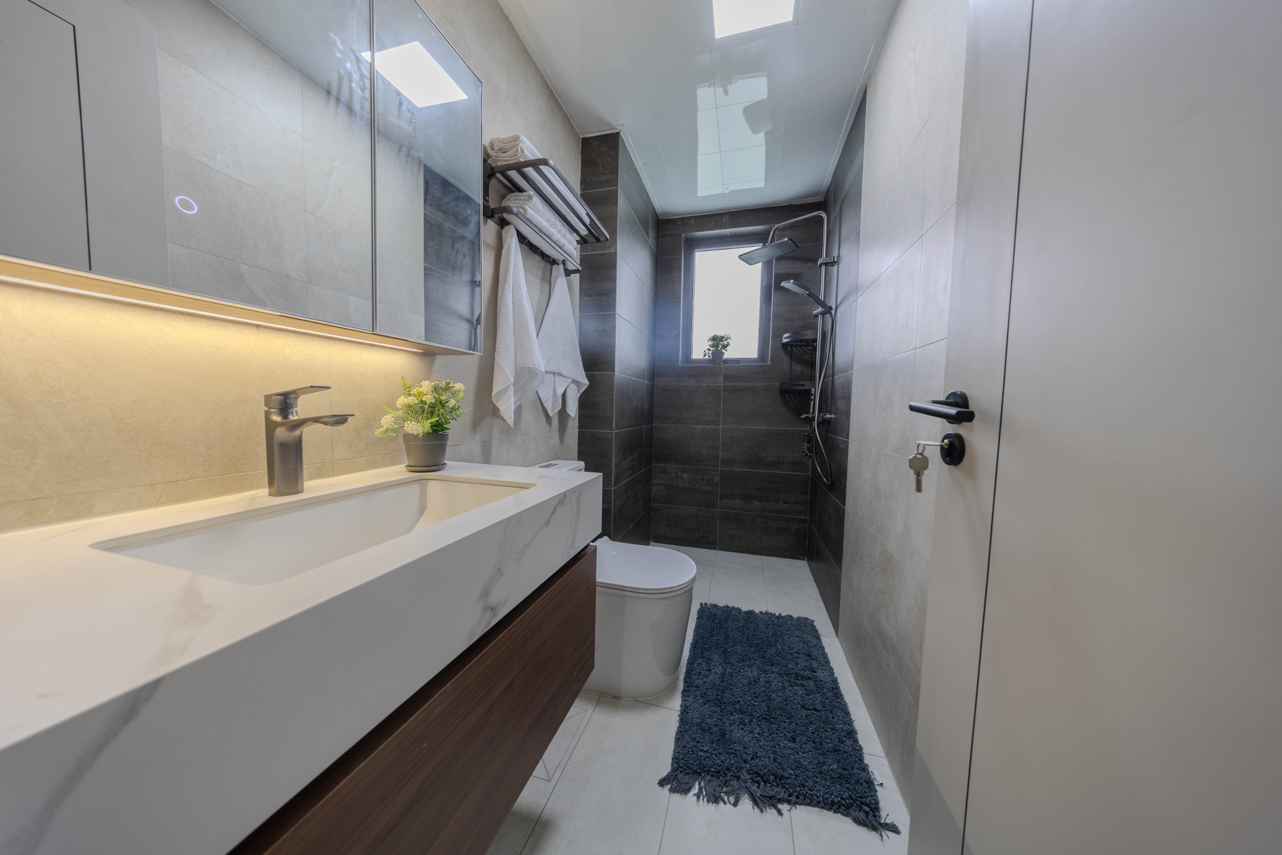 Montbleu apartments modern bathroom featuring a marble-effect vanity sink, illuminated mirror cabinet, rainfall shower, handheld shower, contemporary toilet, and grey tiled walls.
