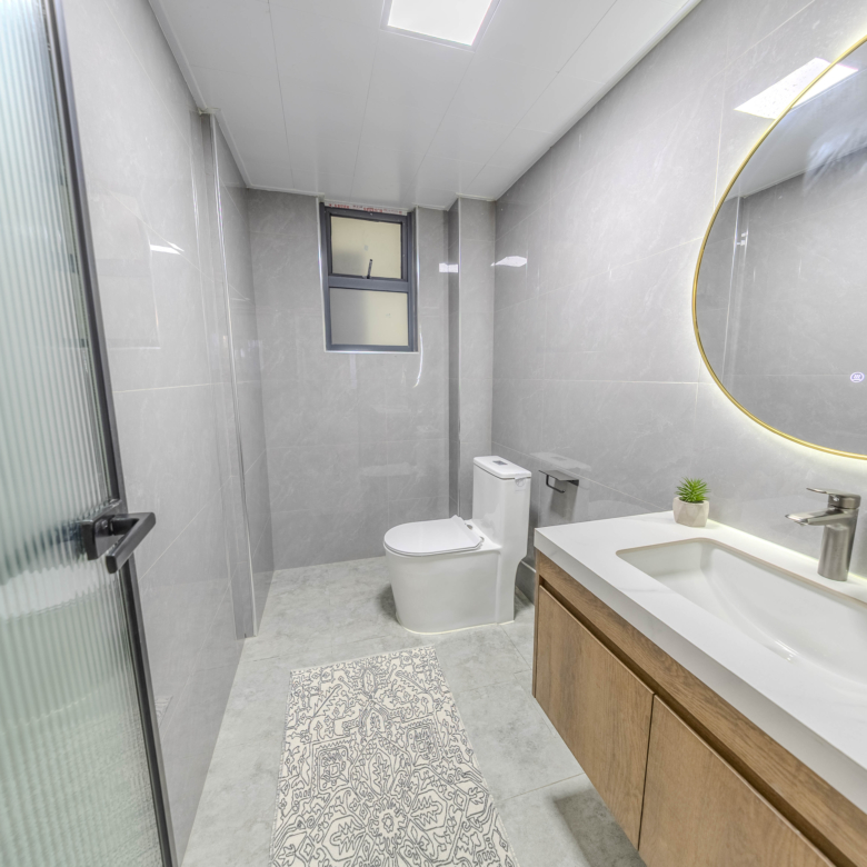 Marina Bay Phase III modern bathroom featuring grey wall tiles, illuminated round mirror, contemporary vanity, and toilet.
