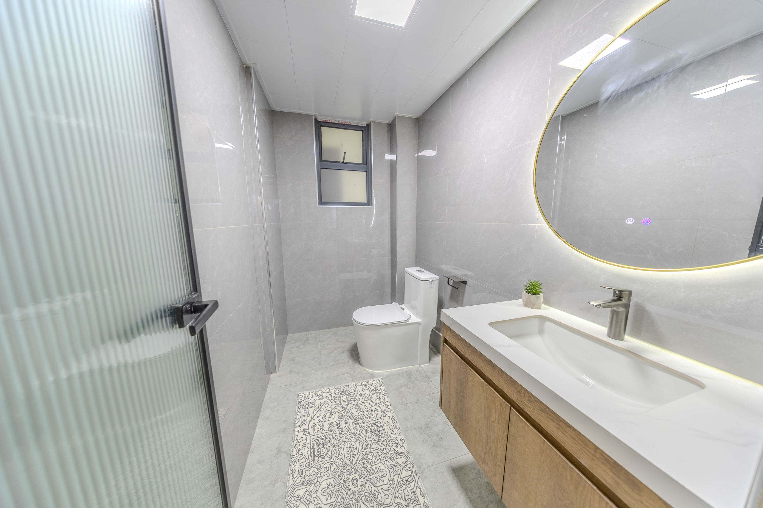 Marina Bay Phase III modern bathroom featuring grey wall tiles, illuminated round mirror, contemporary vanity, and toilet.