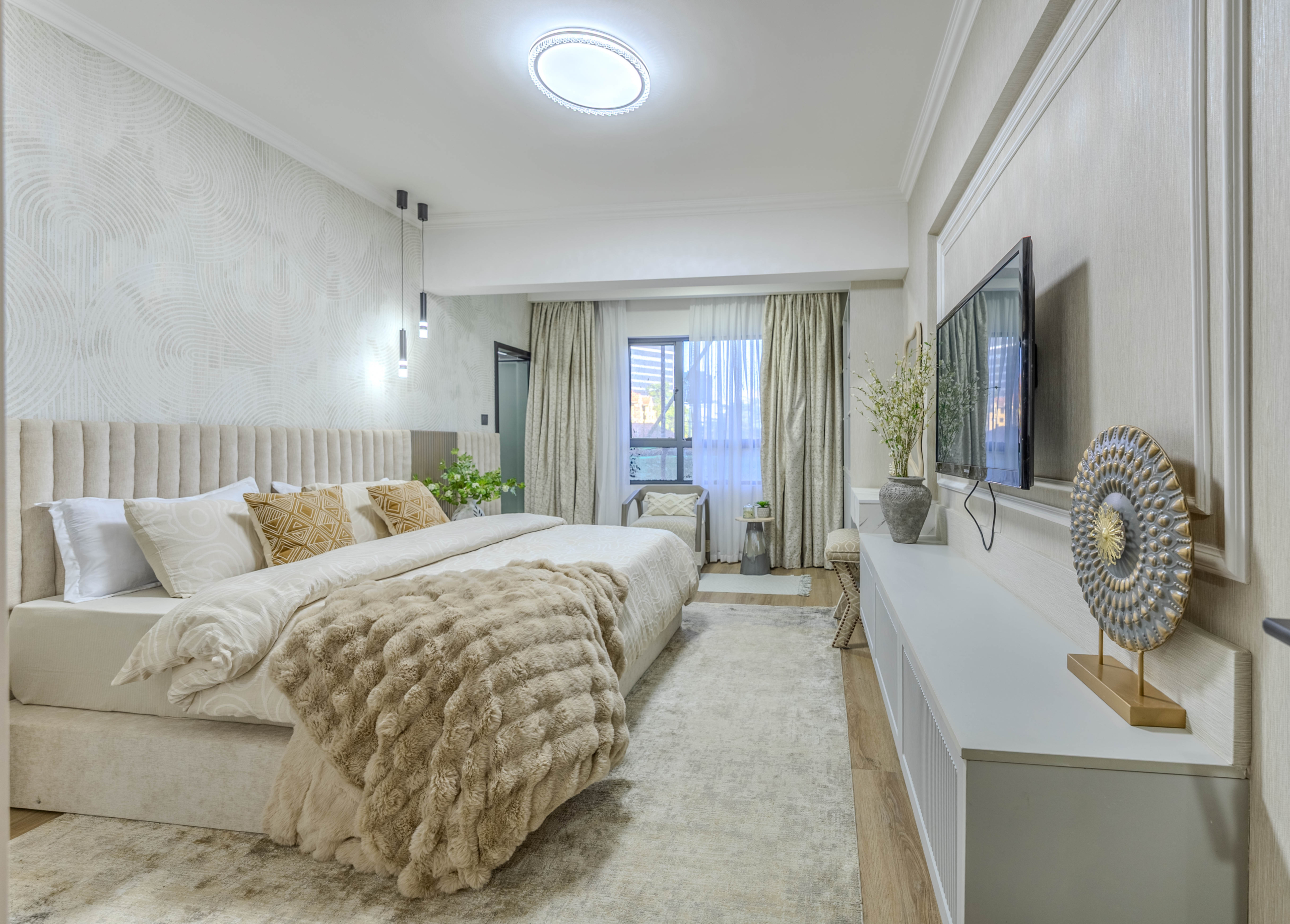 Marina Bay Phase III spacious bedroom with upholstered bed, wall-mounted television, decorative lighting, and large window.