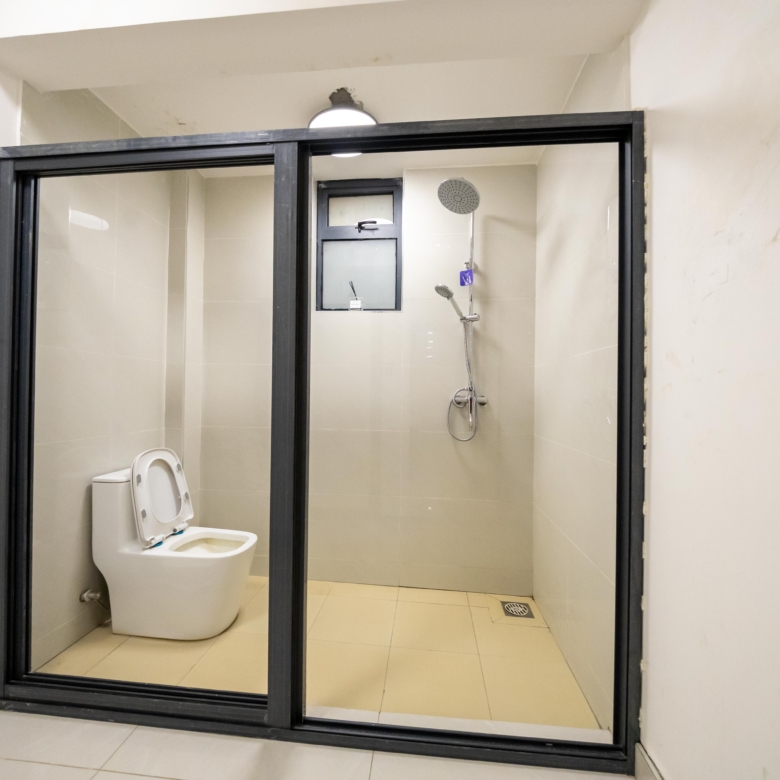 Rhapta One Residency modern bathroom with black-framed glass shower enclosure, white toilet, rainfall shower, and tiled walls.