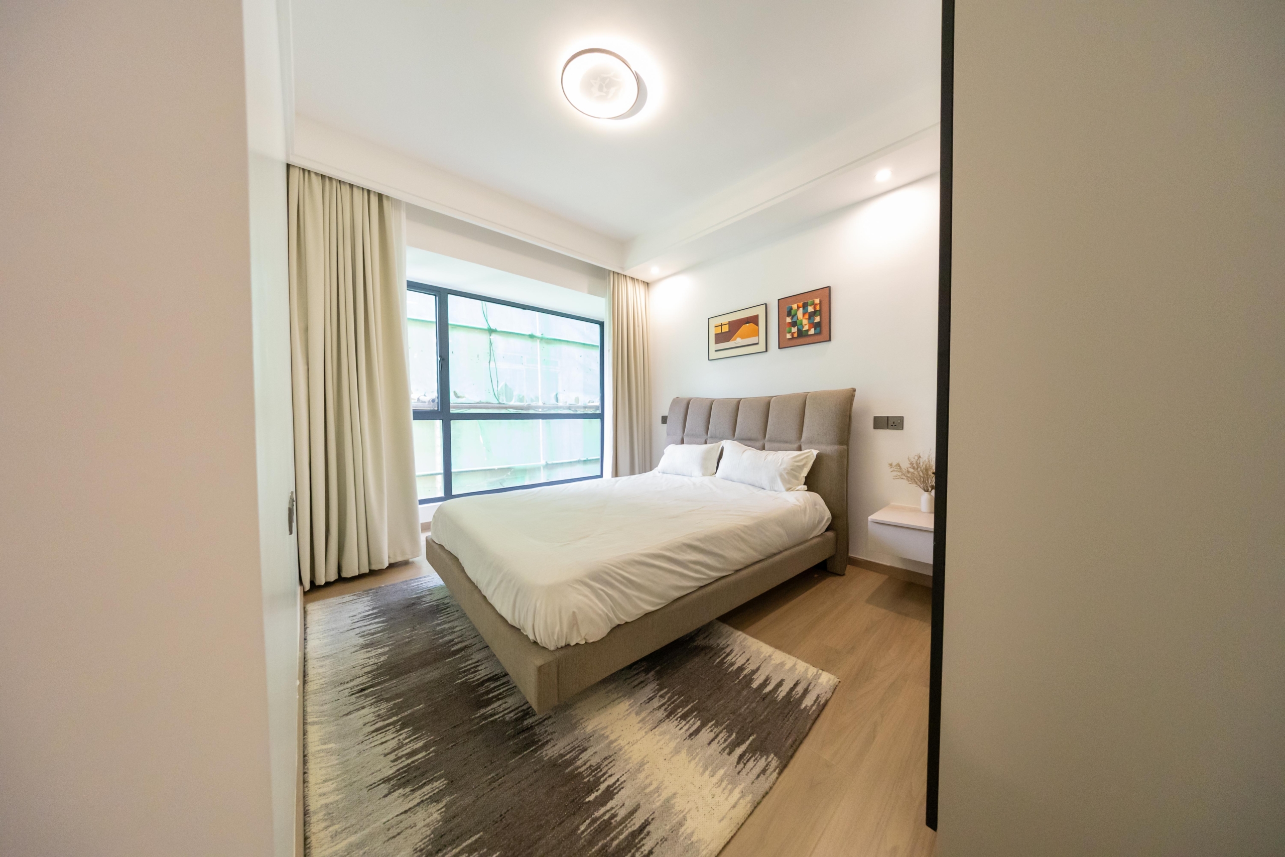 Rhapta One Residency stylish bedroom with upholstered bed, white bedding, large window, floating bedside table, and wooden flooring.