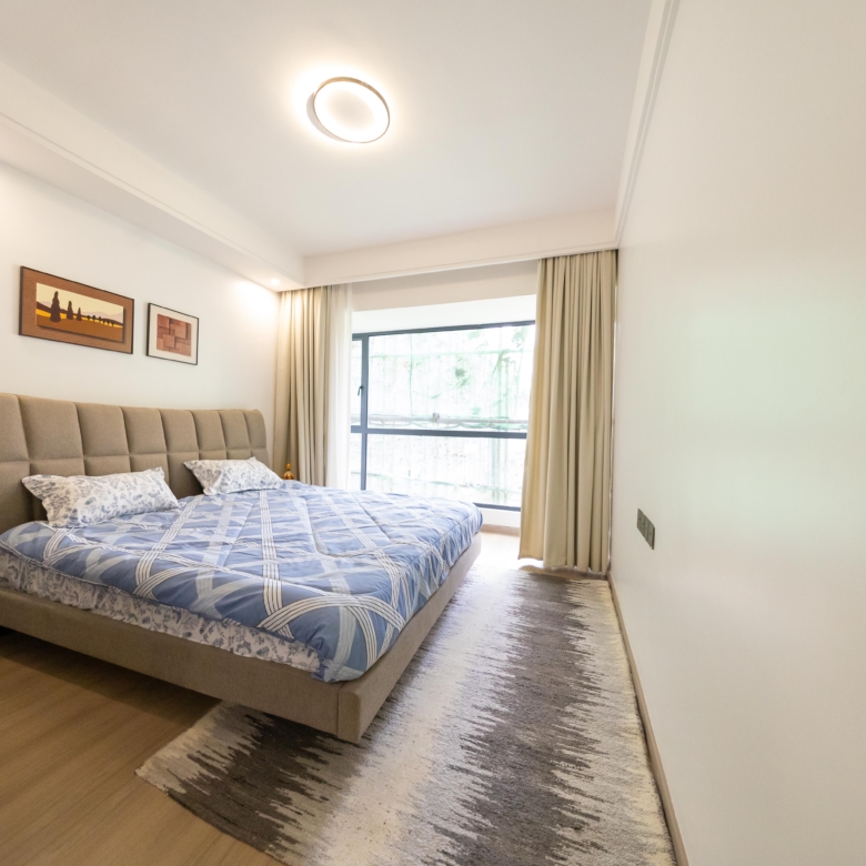 Rhapta One Residency furnished bedroom with upholstered double bed, blue patterned bedding, large window, and modern ceiling lighting.