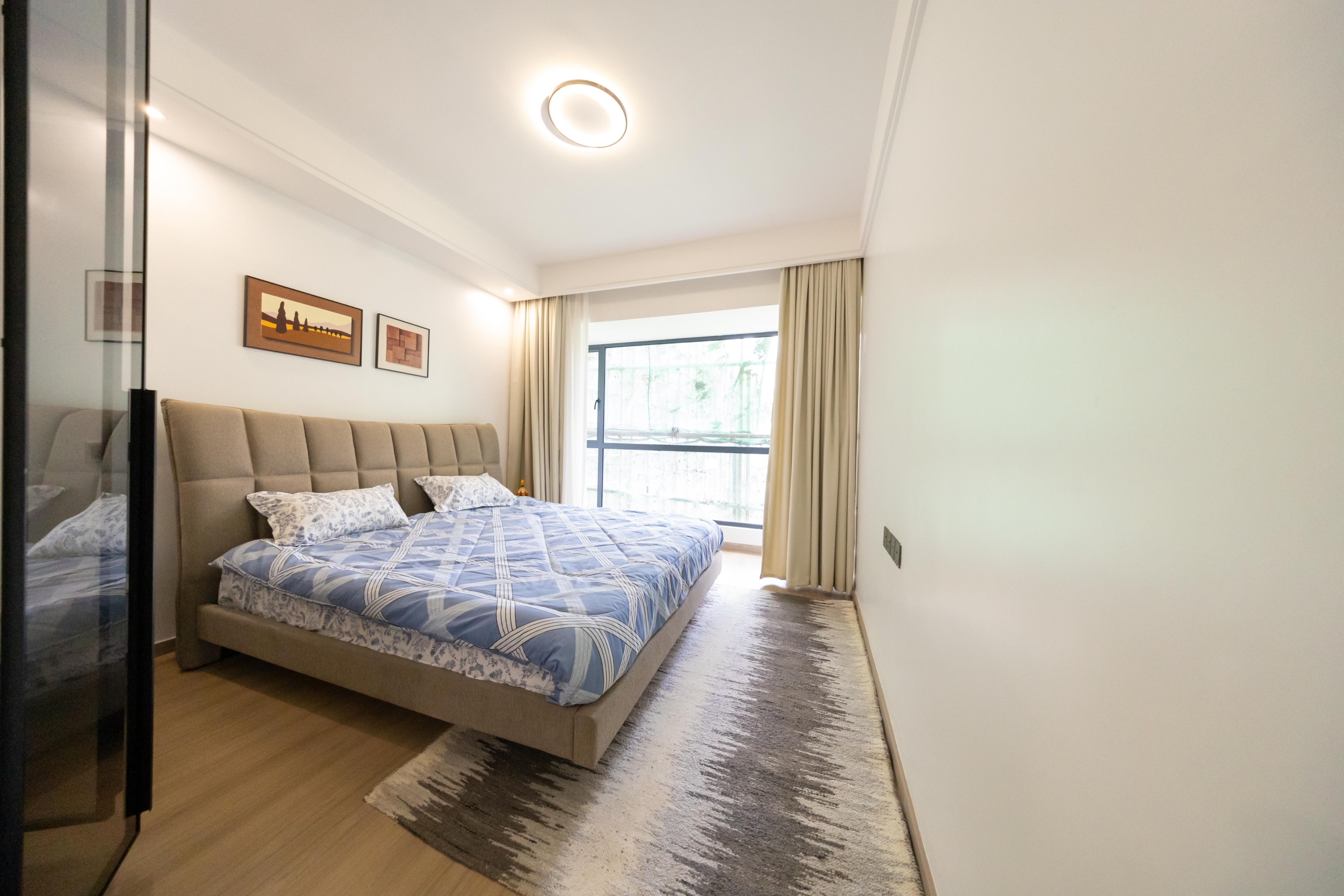 Rhapta One Residency furnished bedroom with upholstered double bed, blue patterned bedding, large window, and modern ceiling lighting.