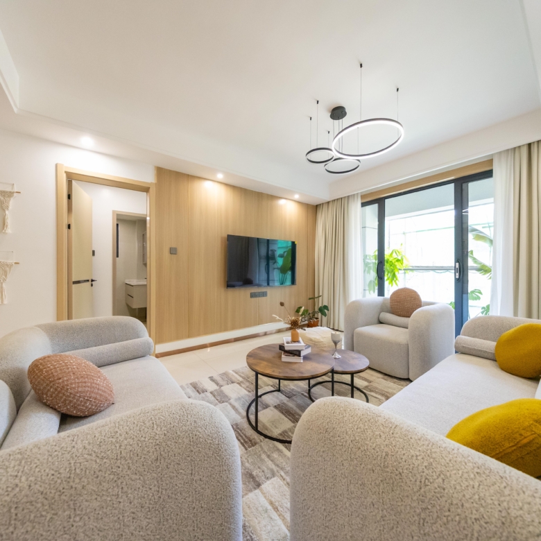 Rhapta One Residency modern living room with cream sofas, wooden TV feature wall, circular coffee tables, and sliding balcony doors.