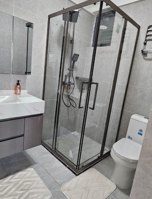 Ilu Green Homes modern apartment bathroom featuring a glass-enclosed shower, rainfall showerhead, handheld shower, vanity sink, wall mirror, toilet, and grey tiled finishes.