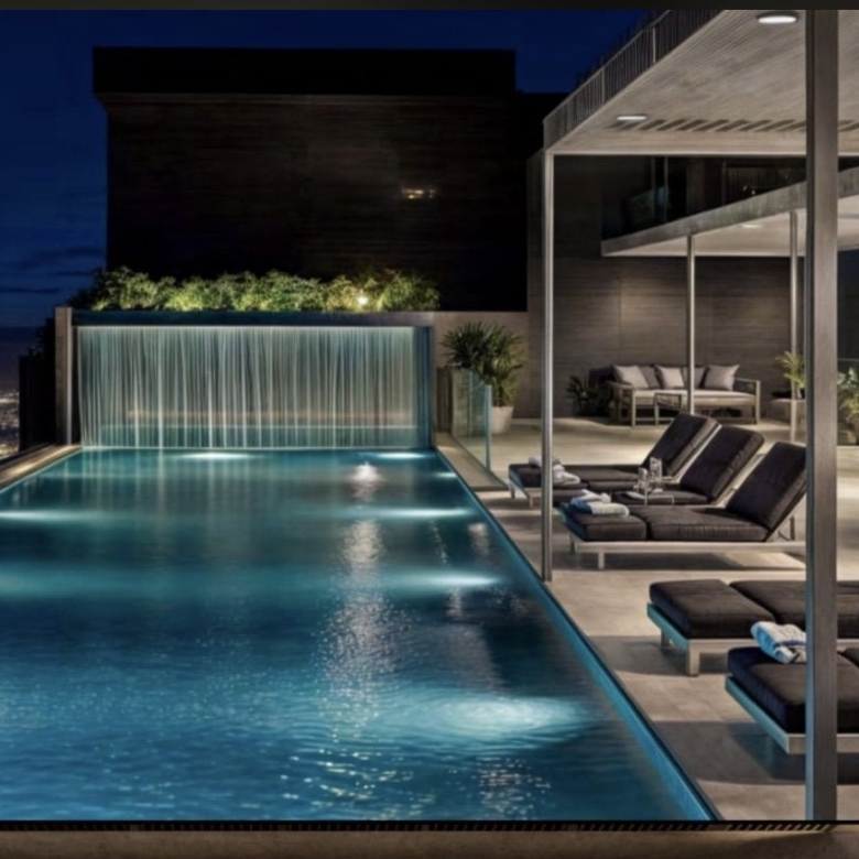 Montbleu apartments rooftop swimming pool rendering featuring an illuminated infinity-style pool, waterfall feature, covered sun loungers, landscaped greenery, and nighttime city views.