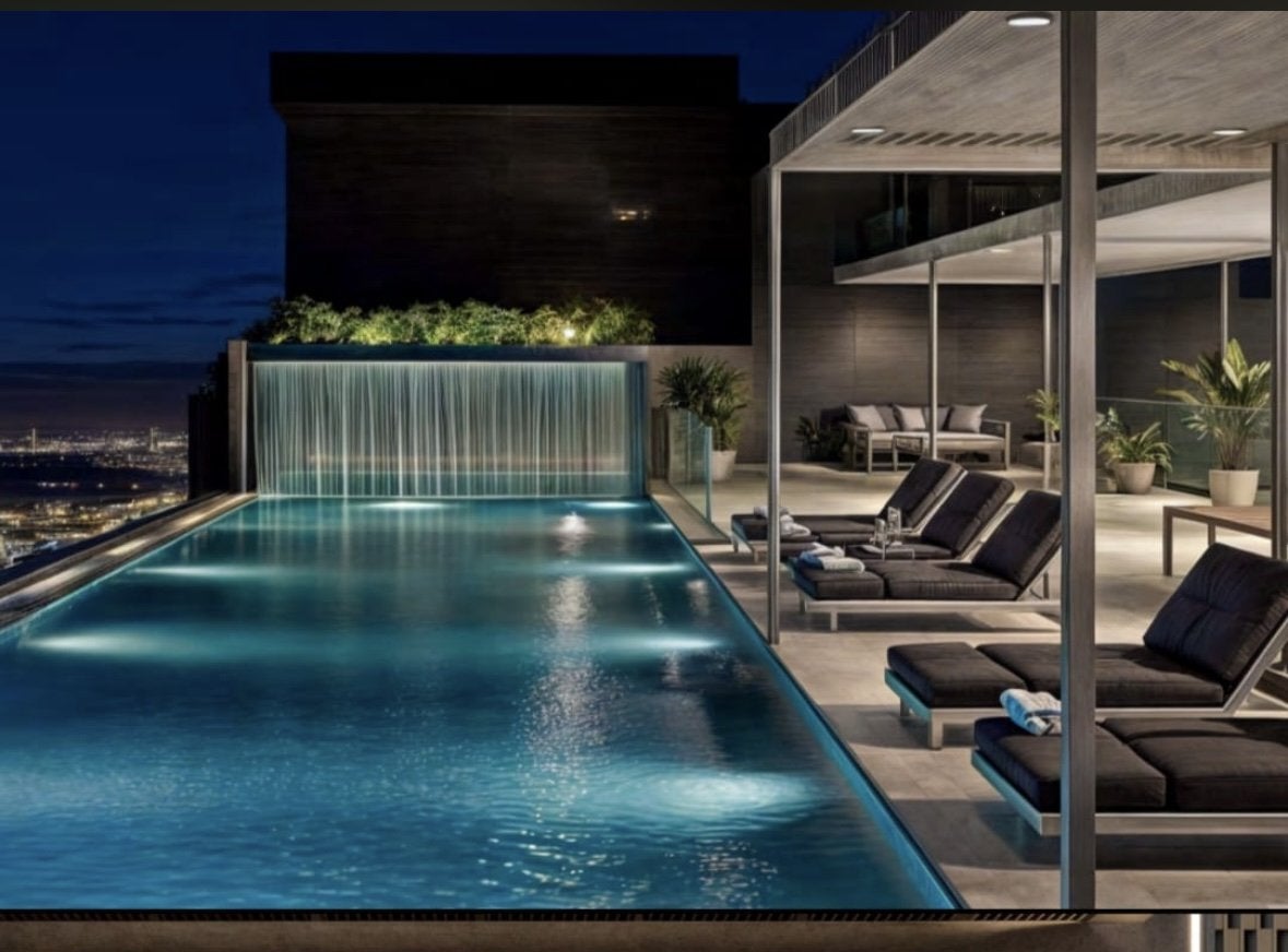 Montbleu apartments rooftop swimming pool rendering featuring an illuminated infinity-style pool, waterfall feature, covered sun loungers, landscaped greenery, and nighttime city views.