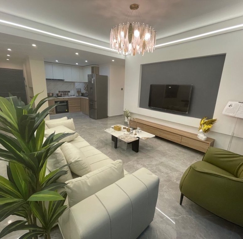 Riara One Residency modern living room with cream sofas, green accent chair, wall-mounted television, chandelier, and open kitchen.