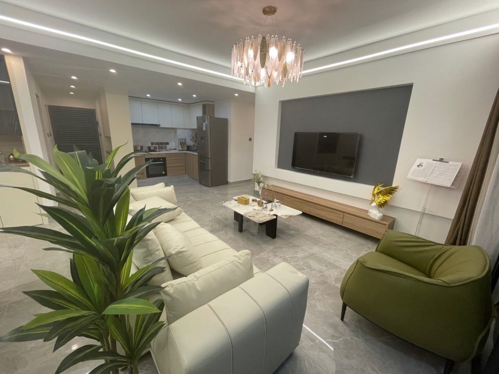 Riara One Residency modern living room with cream sofas, green accent chair, wall-mounted television, chandelier, and open kitchen.