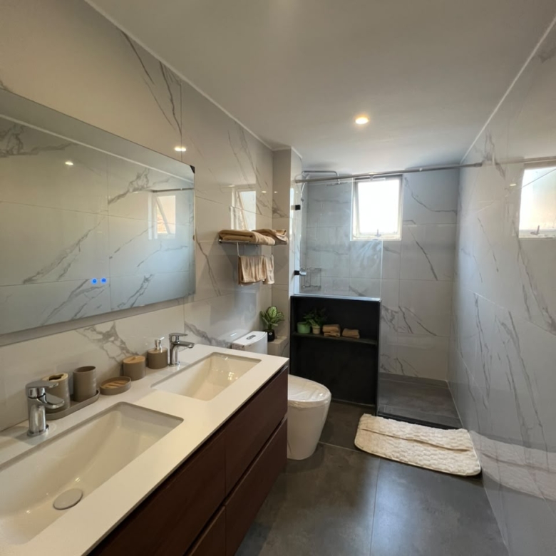 Express View Residency modern bathroom design featuring a glass-enclosed rainfall shower, floating vanity, large mirror, wall-mounted towel rack, and spacious window with blinds.