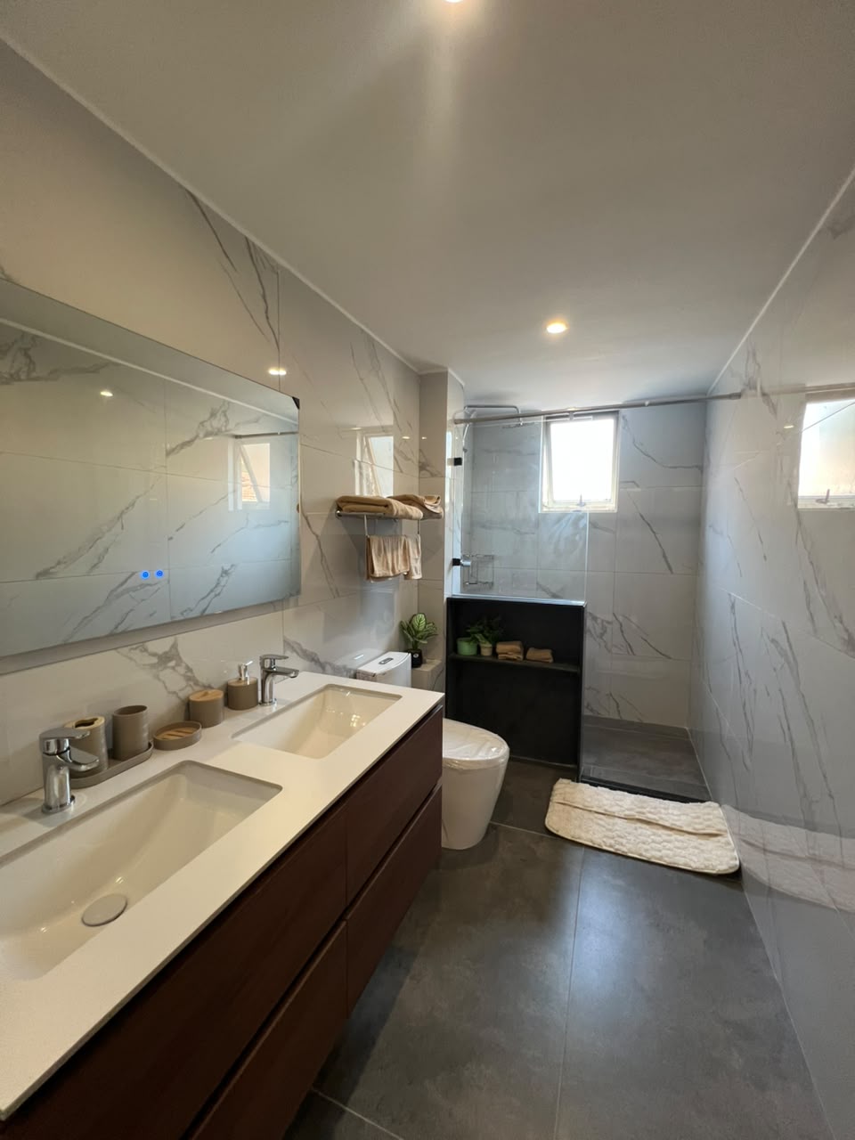 Express View Residency modern bathroom design featuring a glass-enclosed rainfall shower, floating vanity, large mirror, wall-mounted towel rack, and spacious window with blinds.