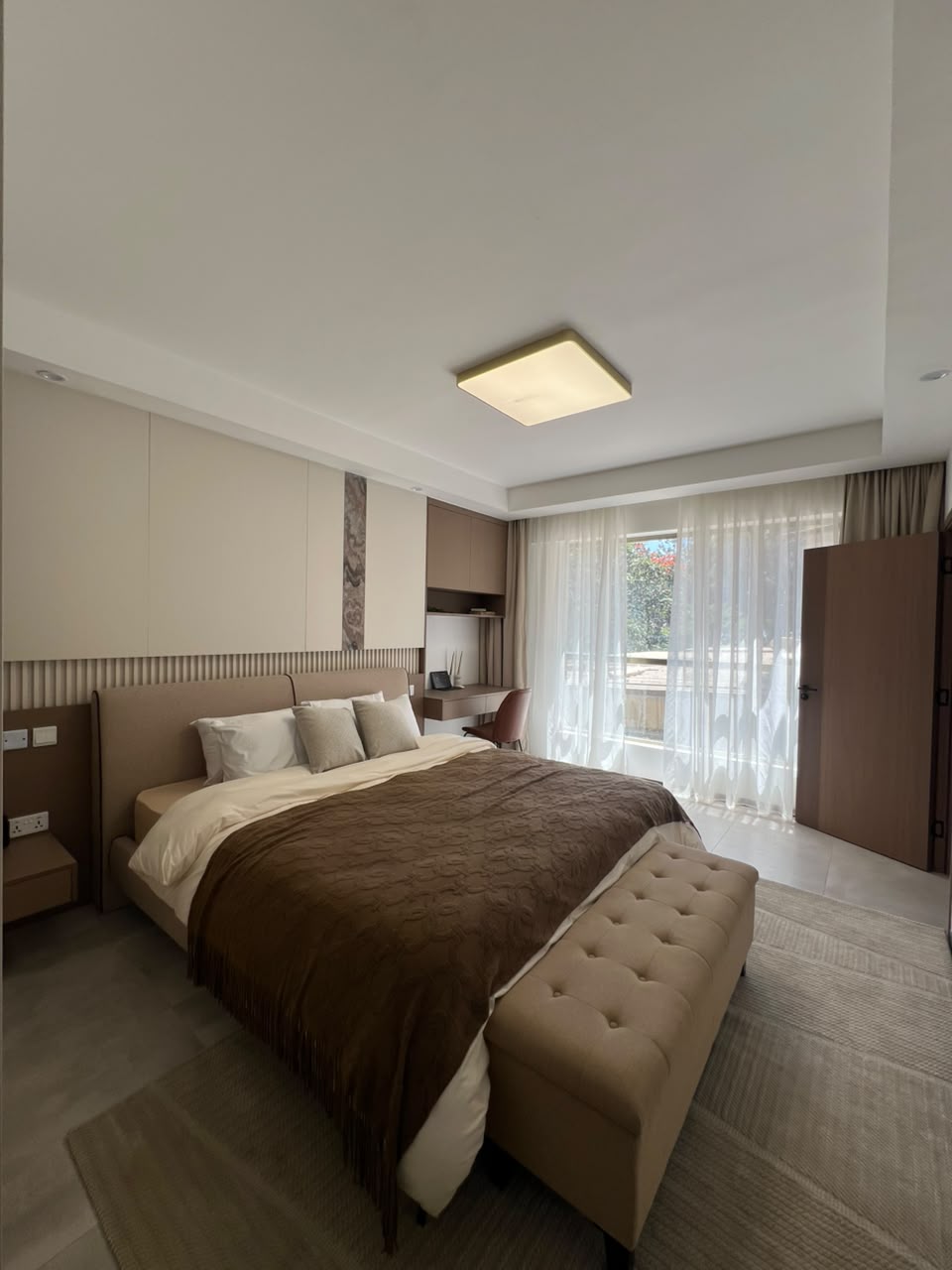 Express View Residency luxury bedroom design featuring a large upholstered bed, fitted wardrobes, modern bedside tables, pendant lights, and a floor-to-ceiling window overlooking the city.