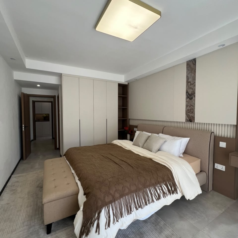 Express View Residency spacious bedroom featuring a large bed, fitted floor-to-ceiling wardrobes, upholstered headboard, decorative wall panels, and a modern ceiling light.