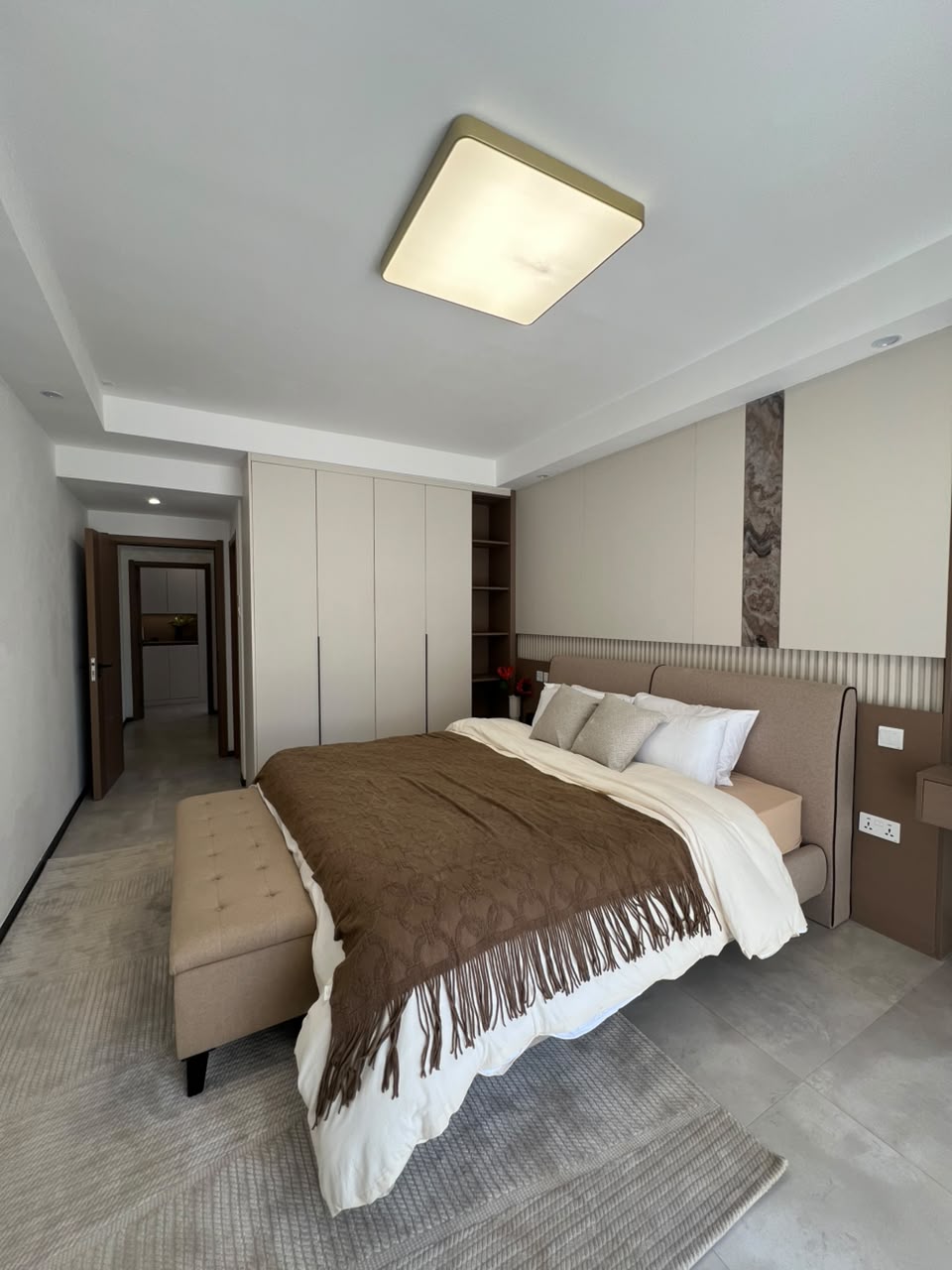 Express View Residency spacious bedroom featuring a large bed, fitted floor-to-ceiling wardrobes, upholstered headboard, decorative wall panels, and a modern ceiling light.