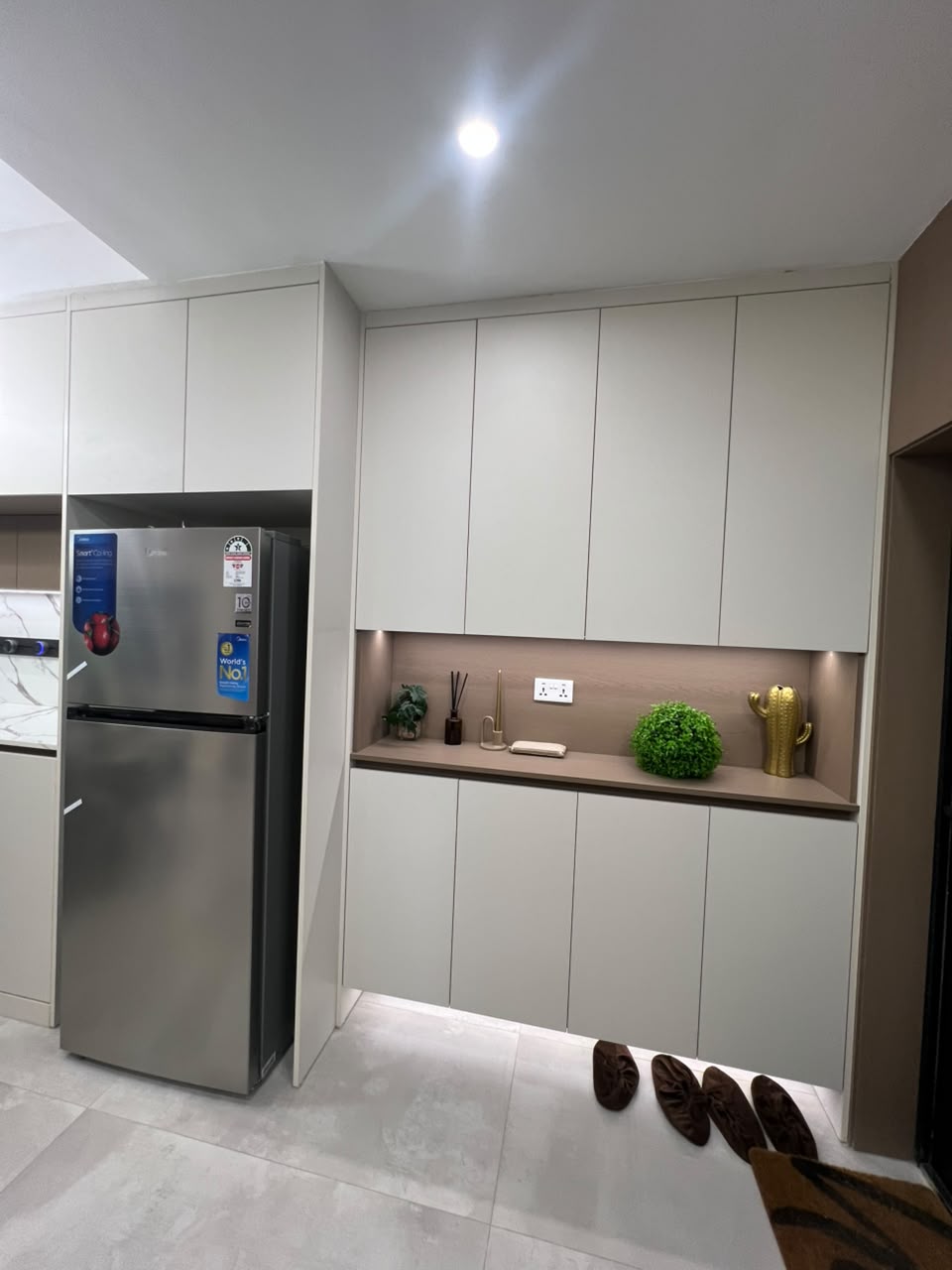 Express View Residency modern kitchen storage area featuring a stainless steel refrigerator, floor-to-ceiling fitted cabinets, illuminated countertop, and decorative open shelving.