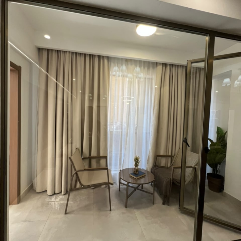 Express View Residency enclosed balcony seating area featuring glass sliding doors, two lounge chairs, a small coffee table, full-length curtains, and modern tiled flooring.