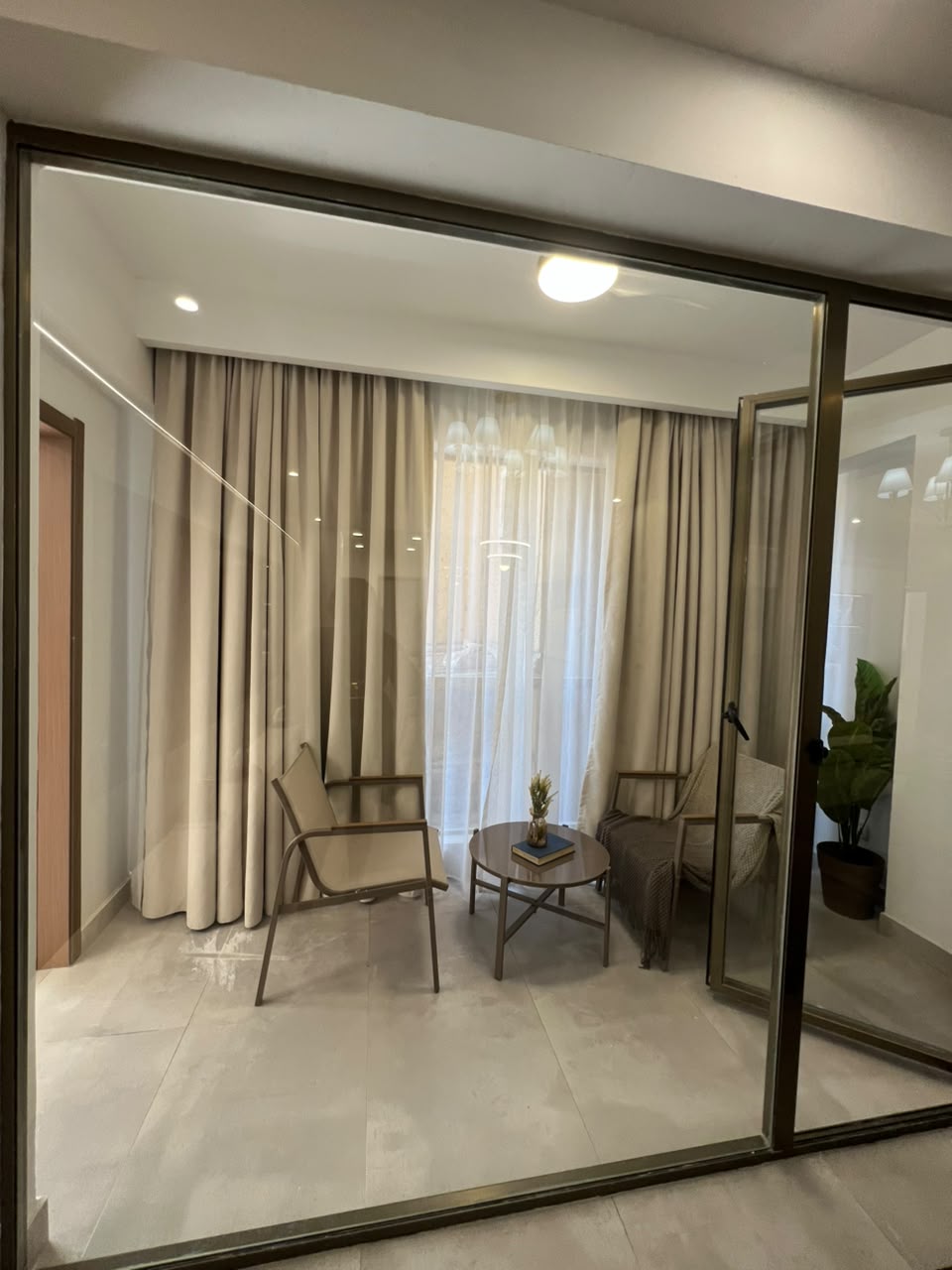 Express View Residency enclosed balcony seating area featuring glass sliding doors, two lounge chairs, a small coffee table, full-length curtains, and modern tiled flooring.