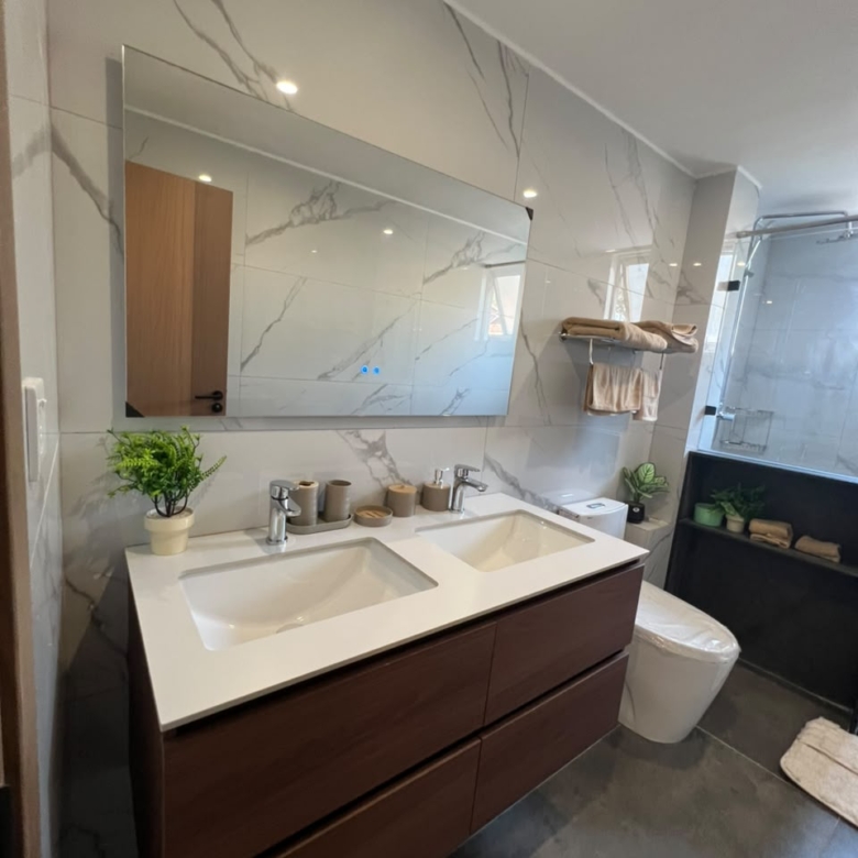 Express View Residency bathroom interior featuring a double-sink vanity, large wall mirror, marble-effect wall tiles, glass-enclosed shower, and contemporary sanitary fittings.