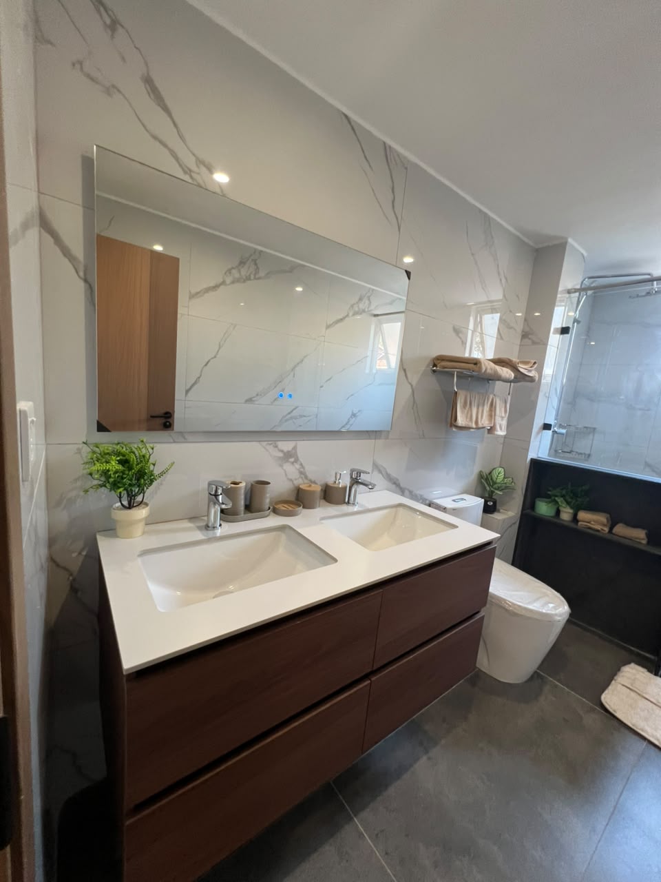 Express View Residency bathroom interior featuring a double-sink vanity, large wall mirror, marble-effect wall tiles, glass-enclosed shower, and contemporary sanitary fittings.