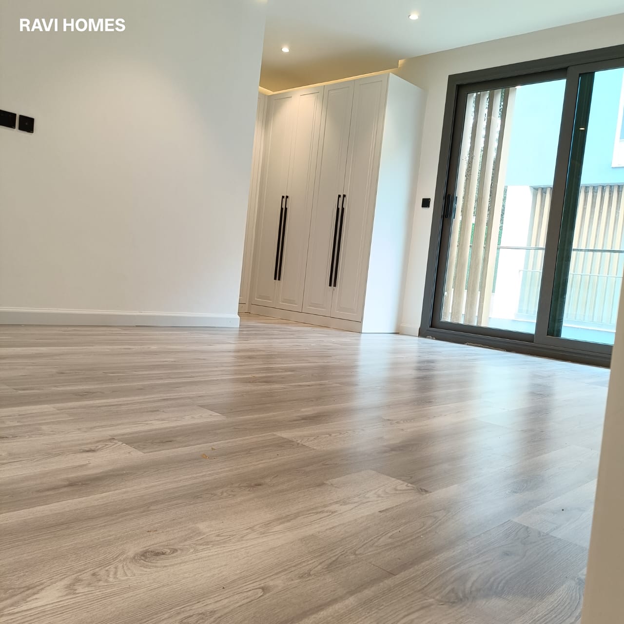 Garden Villas Lavington bedroom with light wood-effect flooring, built-in white wardrobes, neutral walls and black-framed sliding glass doors opening to a balcony.